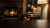 Father and young son reading book together in cozy living room with fireplace. Warm family bonding moment at home during evening storytelling time.