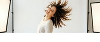 Young African American woman with curly hair dancing joyfully in white sweater by large windows, expressing freedom and happiness in bright indoor space.