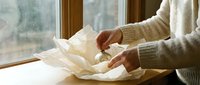 Woman in cozy sweater carefully unwrapping delicate tissue paper package by sunny window, creating warm intimate moment of anticipation and discovery.