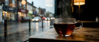 Steaming hot tea cup on wooden table by rainy window with blurred city street lights creating cozy cafe atmosphere for relaxation and warmth.