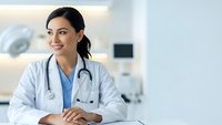 Professional female doctor in white coat with stethoscope sitting at desk in modern medical office, smiling confidently for healthcare consultation.