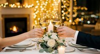 Romantic couple toasting with champagne glasses at elegant dinner table with white roses, candles and fireplace bokeh lights creating intimate atmosphere.