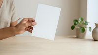 Person with medium skin tone holding blank white paper sheet on wooden desk with potted plant in background for mockup and branding purposes.