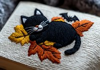 Embroidered black cat sleeping among autumn leaves on textured fabric background, detailed needlework with orange and yellow fall foliage design.