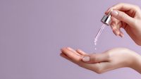Female hands applying skincare serum with dropper against purple background, demonstrating beauty product application technique for cosmetic advertising.