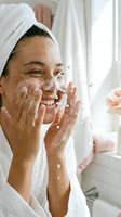 Happy young woman with freckles washing face with foam cleanser in bright bathroom wearing white towel on head after shower morning skincare routine.