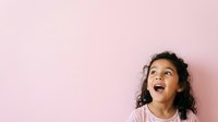 Young girl with dark curly hair and South Asian ethnicity, wearing casual white clothing, expressing joy and amazement with open mouth against soft pink background.