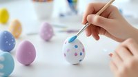 Hand painting polka dots on white Easter egg with pastel colored eggs in background on bright surface, creative holiday craft activity and decoration process.