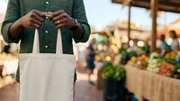 Person holding blank canvas tote bag at farmers market with fresh produce and vendors in blurred background, suitable for eco-friendly shopping and sustainable lifestyle marketing.
