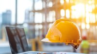 Yellow construction hard hat and laptop on desk with sunlit office window background, representing workplace safety and modern construction management.