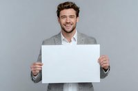 Young businessman in gray suit holding blank white sign with cheerful smile against neutral background, perfect for adding custom message.