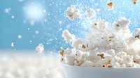 Fresh popcorn explosion with flying kernels against blue background, dramatic food photography capturing motion and texture in bright lighting.
