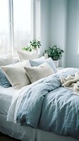Serene bedroom interior with unmade bed featuring soft blue and cream linens, decorative pillows, and potted plants by bright window with sheer curtains.