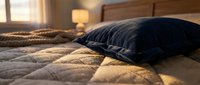 Cozy bedroom scene with dark pillow resting on quilted bedding, warm sunlight streaming through window, creating comfortable home interior ambiance for relaxation and sleep design projects.