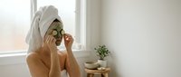 Young woman applying cucumber slices on eyes for natural skincare routine in bright bathroom with towel on head for spa treatment at home.