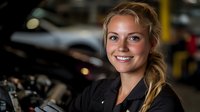 Young female auto mechanic with blonde hair and friendly smile working in repair garage, wearing black uniform, standing near car engine in workshop.