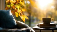 Cozy autumn morning scene with coffee cup on stack of books, blue cushion by window, golden sunlight and fall leaves creating warm atmosphere.