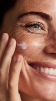 Close-up of woman with freckles applying facial cream, showcasing natural beauty with bright blue eyes and genuine smile.