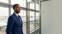 Professional African American businessman in navy suit smiling confidently at modern airport terminal with large windows overlooking aircraft and runway.
