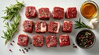 Raw beef tenderloin cut in cubes with fresh rosemary, garlic, peppercorns and olive oil arranged on white background, top view of meat preparation.