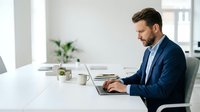 Caucasian male professional in dark blue blazer working on laptop at white desk in modern office with plant and natural lighting.