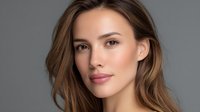 Young Caucasian woman with natural makeup and flowing brown hair poses confidently against gray background, showcasing fresh glowing skin and modern beauty look.