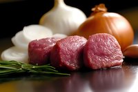 Raw beef tenderloin cubes with fresh garlic, onions and rosemary on wooden surface in natural lighting, ready for gourmet cooking preparation.
