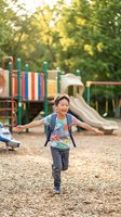 Happy Asian boy running with arms outstretched at colorful playground equipment during golden hour summer day for active childhood and outdoor recreation concepts.