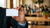 Mature male professional holding blank white business card in modern office environment with blurred colleagues and plants in background.