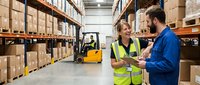 Two warehouse workers in safety vests discussing logistics while forklift operates in background. Industrial teamwork and supply chain management concept.