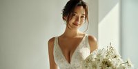 Young Asian bride in elegant white lace wedding dress holding bouquet of white flowers, smiling warmly in soft natural light for bridal photography.