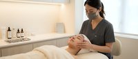 Asian female esthetician in mask performs facial massage on relaxed client in modern spa salon with skincare products on shelf.