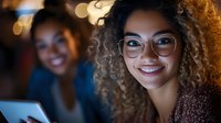 Young African American businesswoman with curly hair and glasses smiling at camera in modern office environment with colleague working in background.
