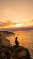 Woman celebrating freedom with raised arms on dramatic coastal cliff during golden sunset over ocean waves for wellness and travel concepts.