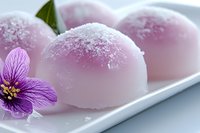 Pink mochi dessert balls dusted with powdered sugar on white plate, decorated with purple flower and green leaf, close up view of traditional Japanese sweets.