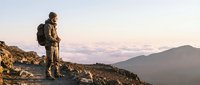 Middle-aged male hiker with backpack standing on rocky mountain ridge at sunset, overlooking misty valleys and distant peaks during outdoor adventure and wilderness exploration.