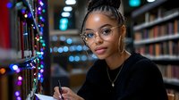 Young African American female student wearing glasses studies in library at night with colorful bokeh lights creating cozy atmosphere while taking notes and smiling at camera.
