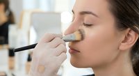 Professional makeup artist applying foundation with brush to young Caucasian woman's face during beauty treatment session in salon studio.