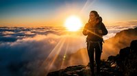 Woman hiker standing on mountain peak above clouds at sunrise, wearing backpack and athletic gear, enjoying scenic golden hour landscape view.