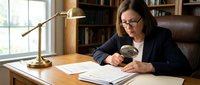 Professional businesswoman with glasses examining documents with magnifying glass at wooden desk in modern office interior with brass lamp and bookshelf background.