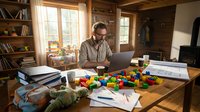 Middle-aged Caucasian male professional working at wooden desk with laptop, surrounded by colorful building blocks, papers, and office supplies in warm wooden home office with bookshelves and natural light.
