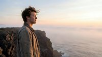 Young man with curly hair standing on coastal cliff overlooking ocean at sunset, contemplating future with peaceful expression and warm golden light.