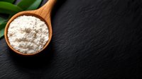 White flour in wooden spoon on black stone background with green leaf decoration, top view. Food ingredients and cooking concept for bakery and culinary designs.