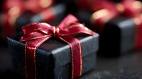 Black gift box with elegant red satin ribbon bow on dark background with bokeh lights. Luxury present packaging for special occasions and holidays.