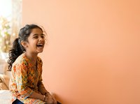 Young South Asian girl with dark wavy hair laughing joyfully against warm peach background, wearing patterned orange shirt, expressing happiness and childhood joy for lifestyle and family content.