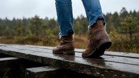 Legs in jeans and brown leather boots standing on a wet wooden boardwalk in a rainy forest landscape, outdoor adventure concept.