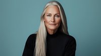 Mature woman with long silver hair wearing black turtleneck sweater against teal background, expressing confidence and wisdom in professional studio portrait.