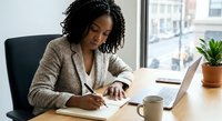 Professional African American businesswoman writing notes at modern office desk with laptop and coffee cup near window.
