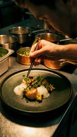 Chef plating gourmet dish with tweezers, adding microgreens and sauce to seared protein with puree on gray ceramic plate in professional kitchen.