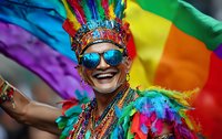 Joyful participant wearing vibrant rainbow costume, feathers, and blue reflective sunglasses celebrates at pride parade against colorful flag background.